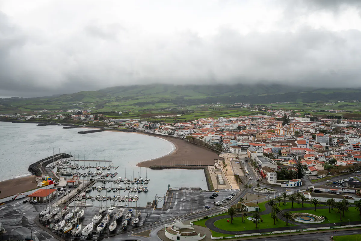 An aerial view of Praia da Vitória on Terceira Island shows a curved sandy bay, with a marina full of sailboats on one side and a town of white houses with orange rooftops on the other, and green hills hidden behind thick clouds in the background.
