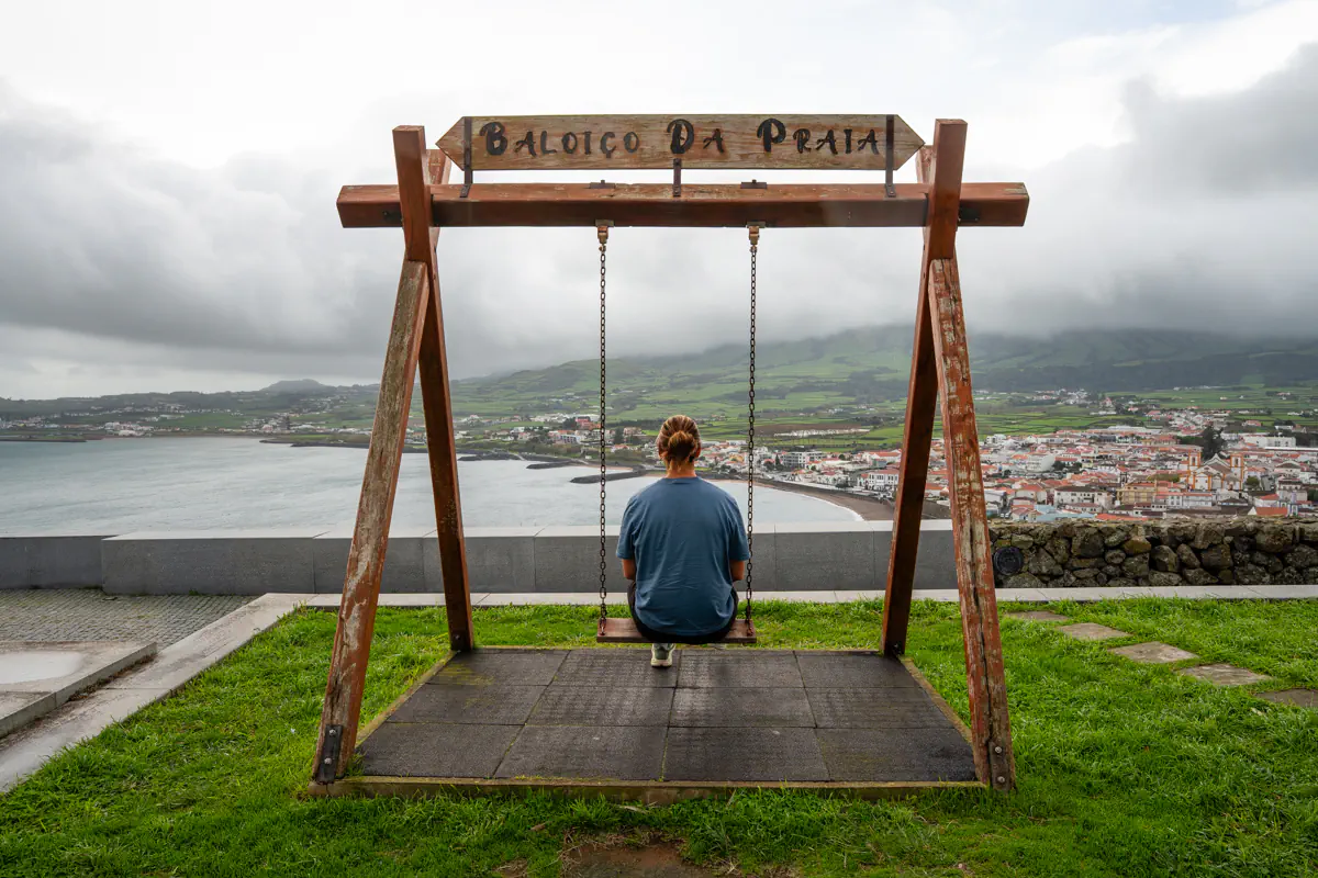 A person sits on a wooden chain swing at a hilltop viewpoint on Terceira Island, looking out over the coastline and terracotta rooftops of Praia da Vitória below, with a sign above reading "Baloiço Da Praia" and green hills rolling into the misty distance.
