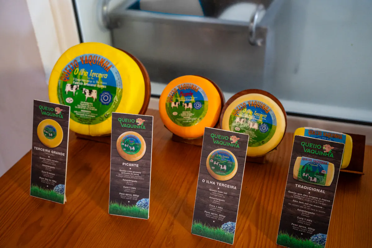 Three wheels of Queijo Vaquinha cheese in yellow, orange, and blue-labelled wax sit on a wooden counter alongside four product cards. The cheeses are "Terceira Grande," "Picante," "O Ilha Terceira," and "Tradicional."
