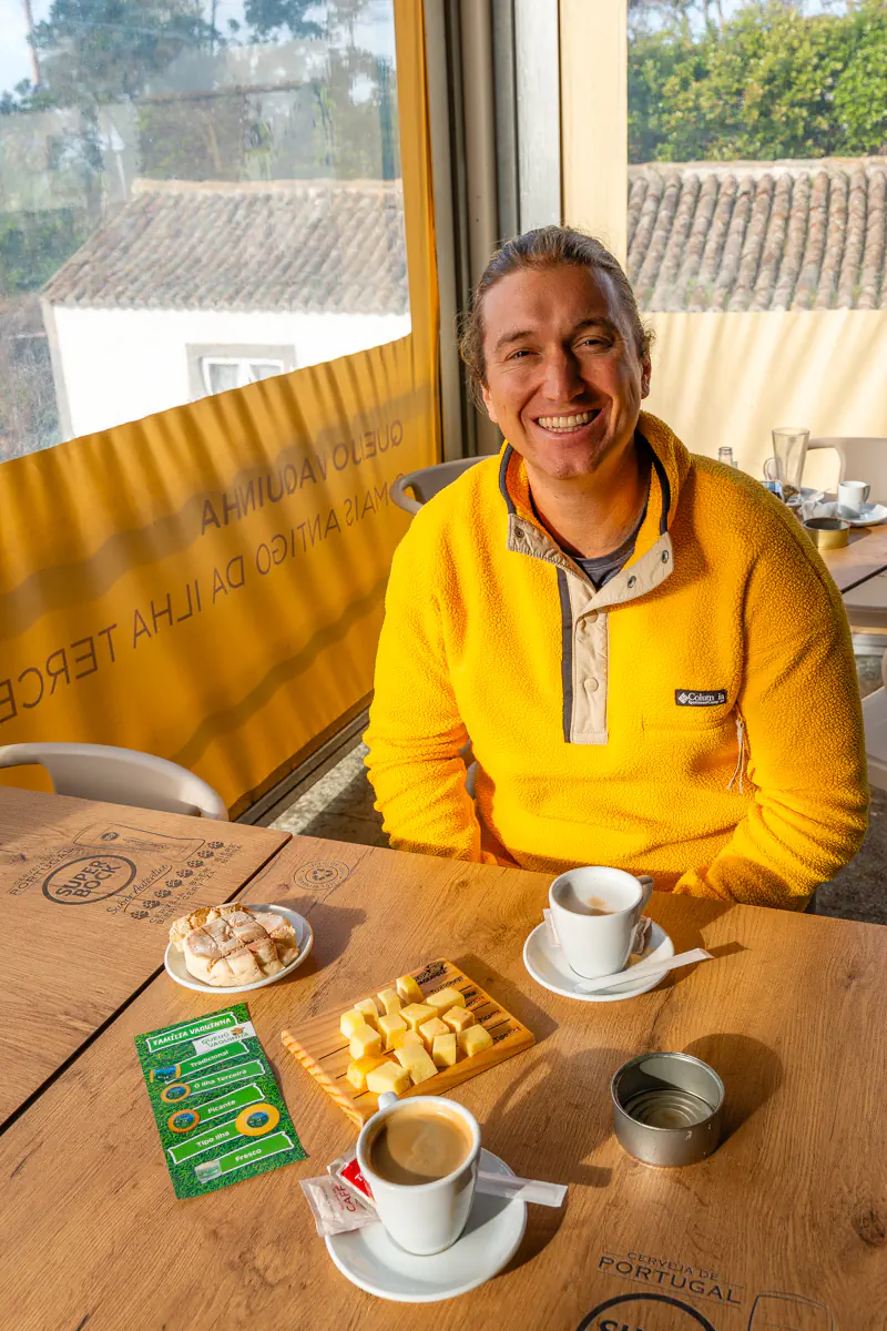 A smiling person sits at a wooden table inside the Queijo Vaquinha shop on Terceira Island, with two coffees, a bread roll, and a board of cubed cheese samples in front of them, and a yellow branded banner reading "Queijo Vaquinha Mais Antigo Da Ilha Terceira" visible behind them.