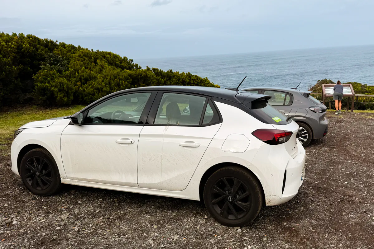 Two rental cars are parked on a gravel pullout at a coastal viewpoint on Terceira Island, with a person reading an information board beside the Atlantic Ocean and dense green shrubs framing the scene under an overcast sky.