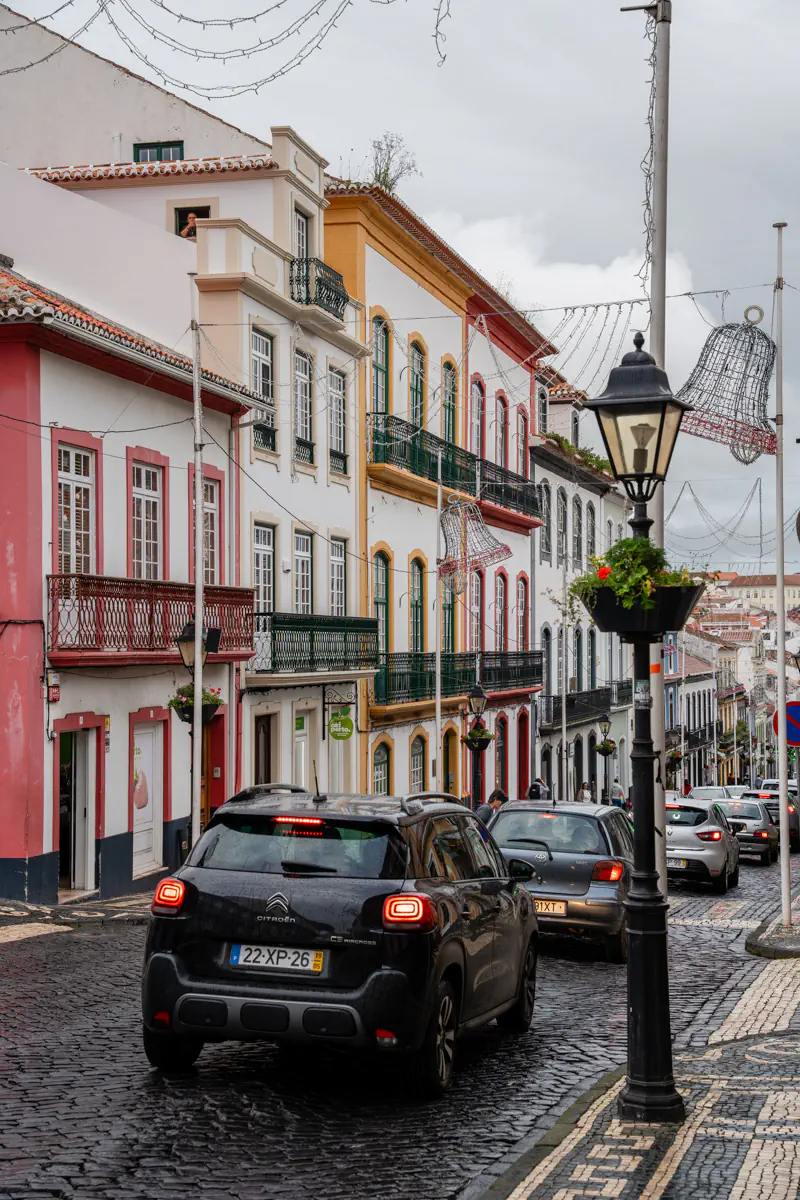 A wet cobblestone street in Angra do Heroísmo on Terceira Island is lined with colourful buildings with balconies. There are Christmas light decorations strung overhead and a black Citroën SUV stopped in traffic under a grey sky.
