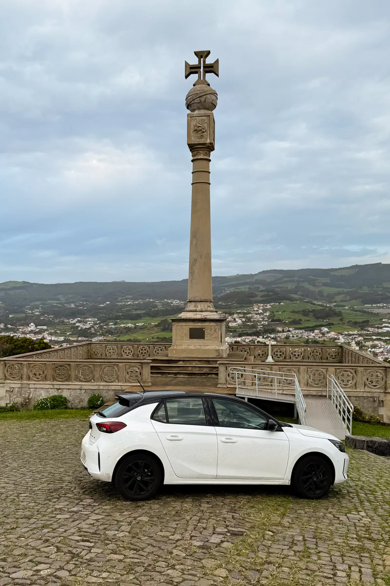 A white rental car is parked on cobblestones beside a tall stone pillar monument topped with a cross at a hilltop viewpoint on Terceira Island, with a panoramic view of a town and green volcanic hills stretching out behind it under a cloudy sky.