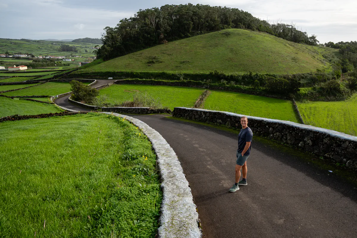 A person stands smiling on a narrow winding road on Terceira Island, surrounded by vivid green fields divided by traditional lava stone walls, with a rounded volcanic cone blanketed in grass and trees rising dramatically behind them.