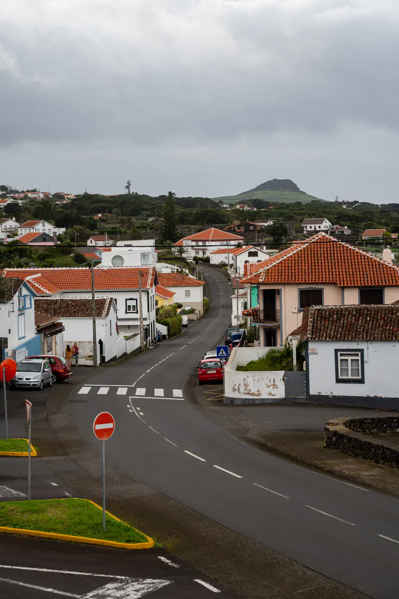 A quiet road winds through a small village on Terceira Island, passing whitewashed houses with terracotta rooftops, a red stop sign at an intersection in the foreground, and a distinctive green volcanic peak rising above the treeline in the distance.