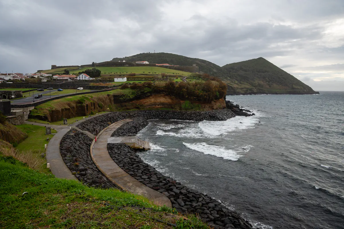 A curved walkway winds along the black rocky shoreline of Angra do Heroísmo on Terceira Island, with waves crashing against the rocks, a green hilltop fort visible above.