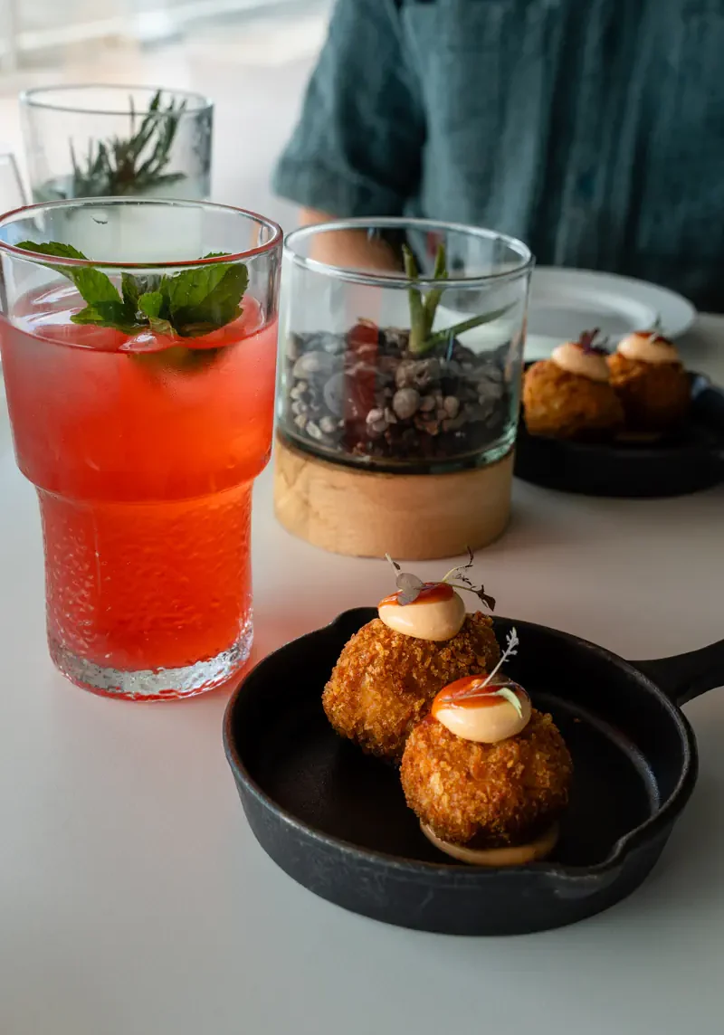 A pink lemonade garnished with fresh mint sits beside two golden-brown croquettes topped with a creamy mayonnaise in a small black pan. The dishes are placed on a light-coloured table with minimalist decor in the background.