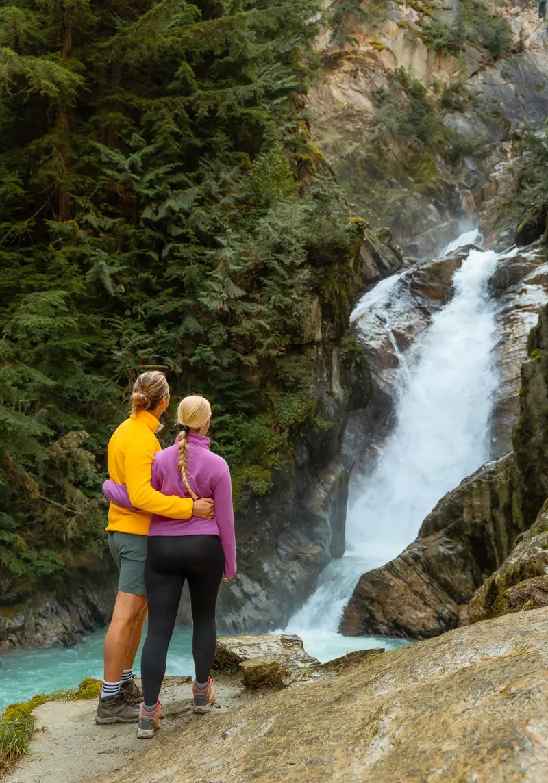 A couple stands arm in arm on a rocky ledge, looking at a waterfall cascading through a forest. The man wears a bright yellow sweater and the woman a pink one, both dressed in hiking gear, surrounded by lush evergreen trees and turquoise water below.