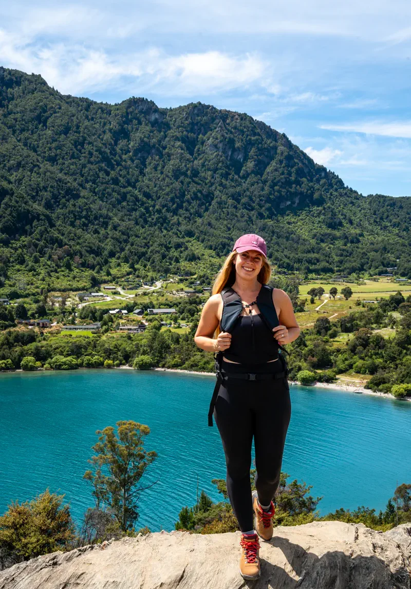 A woman stands on a rocky ledge overlooking a bright blue lake with a small beach and forested hills behind her. The clear water and sunny conditions emphasize the vibrant outdoor setting.