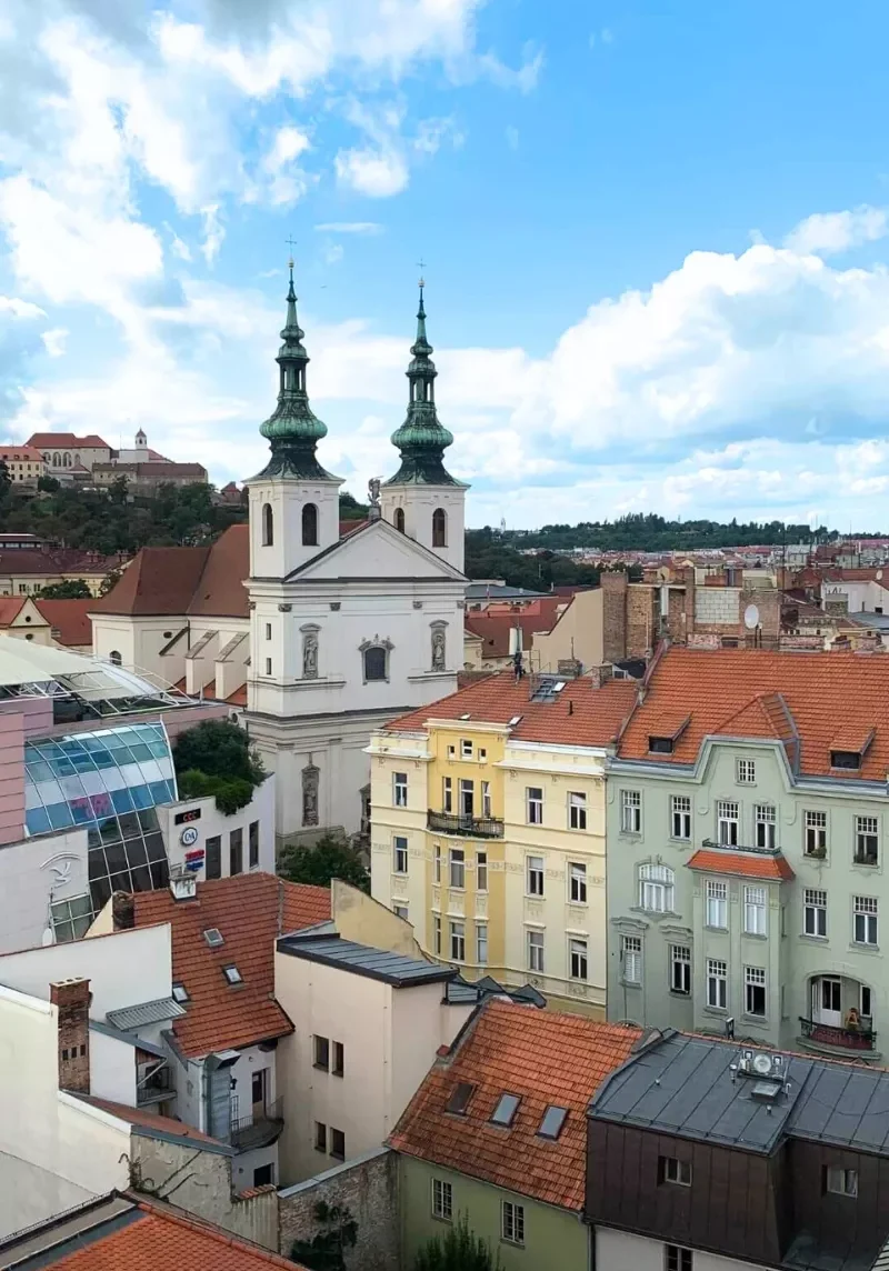 Aerial view over the rooftops of Brno, Czech Republic, featuring the white facade and green spires of the Church of St. Thomas, surrounded by colorful historic buildings and modern glass architecture.