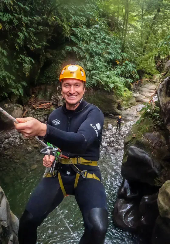 A man in a wetsuit and helmet rappels down a narrow canyon using a safety rope. A rocky stream and green vegetation are visible below him.