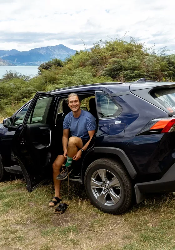 Person in a blue T-shirt sits on the open rear door sill of a dark blue Toyota RAV4 rental, adjusting a sandal on a grassy roadside turnout near Queenstown, New Zealand. Behind the car, Lake Wakatipu and rugged mountains frame the lush, fern-covered landscape.