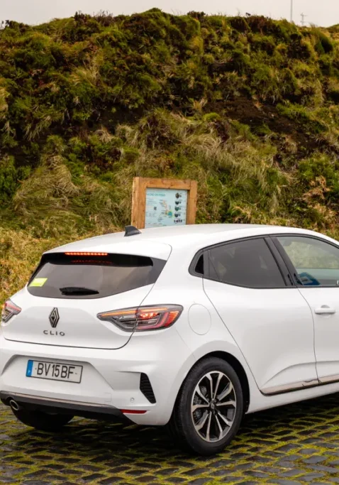 A white rental car is parked on a cobblestone pull-off beside a grassy hillside on São Miguel Island. A small roadside signboard and sloping green terrain surround the vehicle.