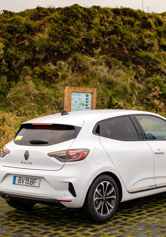 A white rental car is parked on a cobblestone pull-off beside a grassy hillside on São Miguel Island. A small roadside signboard and sloping green terrain surround the vehicle.