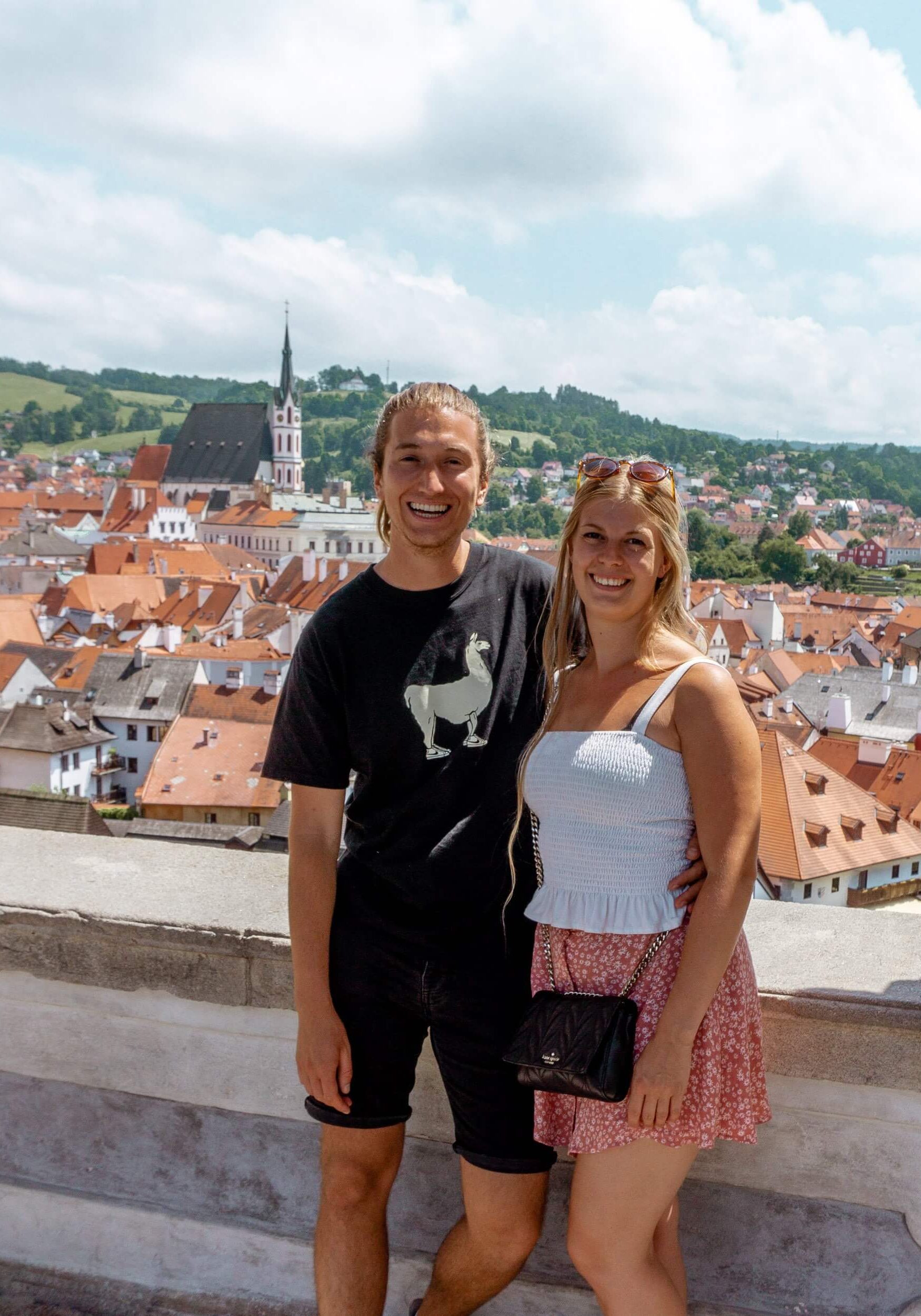 Couple standing above the town center of Cesky Krumlov. You can see all the red rooftops as well as the Church of St. Jakub Vetsi.