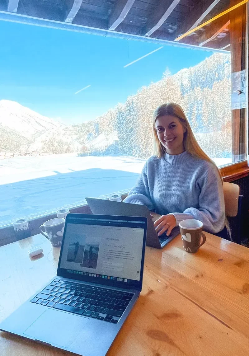A woman in a cozy sweater works on her laptop at a wooden table inside a coliving in Switzerland, with snowy mountains and pine trees visible through the large window behind her. A second open laptop and two mugs of coffee are on the table.
