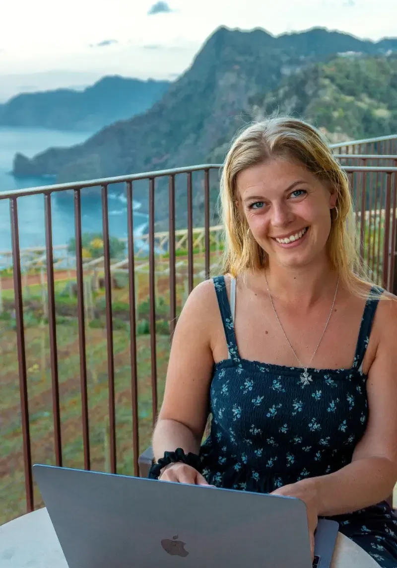 A woman smiles while working on a laptop at an outdoor table, with dramatic coastal cliffs and the ocean in the background. The image captures a relaxed digital nomad moment, blending remote work with a scenic seaside view.