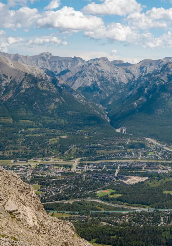 View from the summit of Ha Ling Peak overlooking the town of Canmore, with rows of houses, roads, and the Bow River visible far below. In the distance, rugged peaks of the Fairholme Range rise under a partly cloudy blue sky, creating a beautiful view across the Bow Valley.