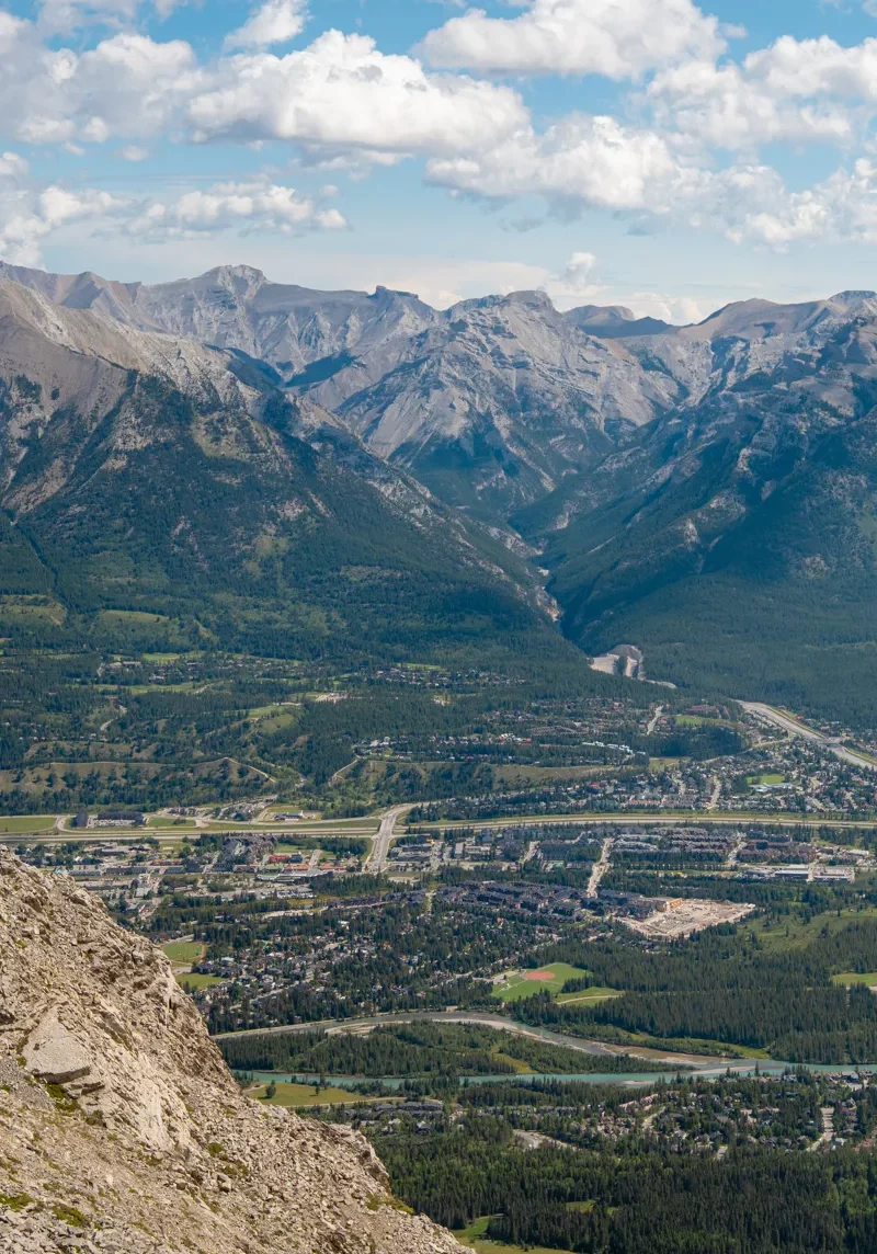 View from the summit of Ha Ling Peak overlooking the town of Canmore, with rows of houses, roads, and the Bow River visible far below. In the distance, rugged peaks of the Fairholme Range rise under a partly cloudy blue sky, creating a beautiful view across the Bow Valley.