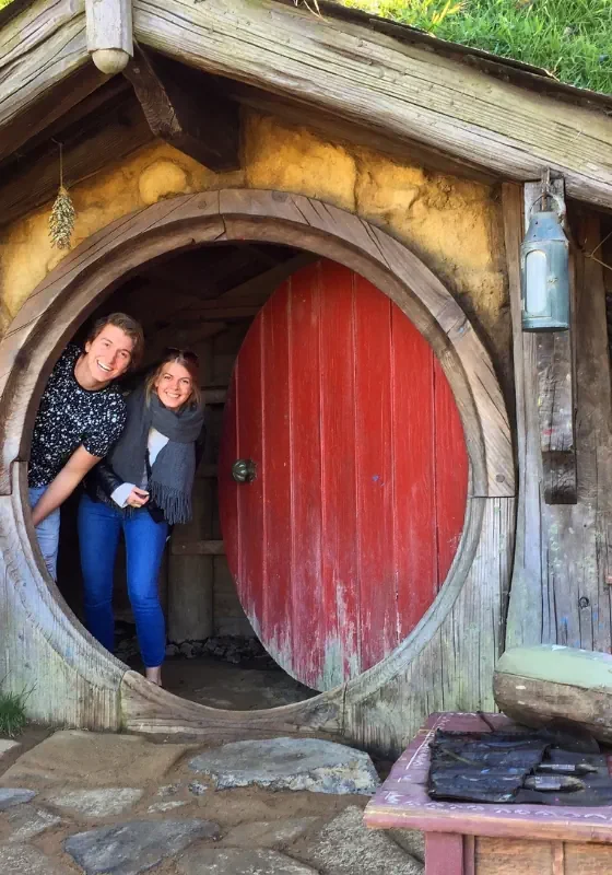 Two people smile while standing in the round doorway of a hobbit house with a red door and rustic wooden beams. Shelves with jars and tools sit beside the entrance, adding to the cozy lived in feel.
