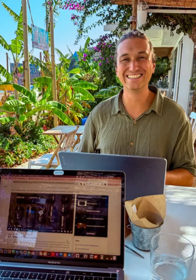 A smiling man works on his laptop at an outdoor café surrounded by banana plants and tropical greenery in Rhodes, Greece. A second laptop in the foreground shows a browser open, with a glass of water on the table.