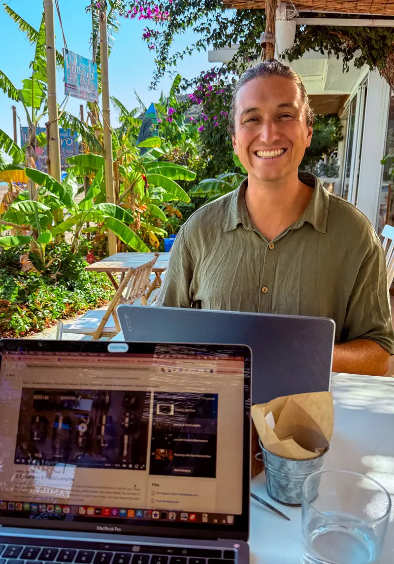 A smiling man works on his laptop at an outdoor café surrounded by banana plants and tropical greenery in Rhodes, Greece. A second laptop in the foreground shows a browser open, with a glass of water on the table.