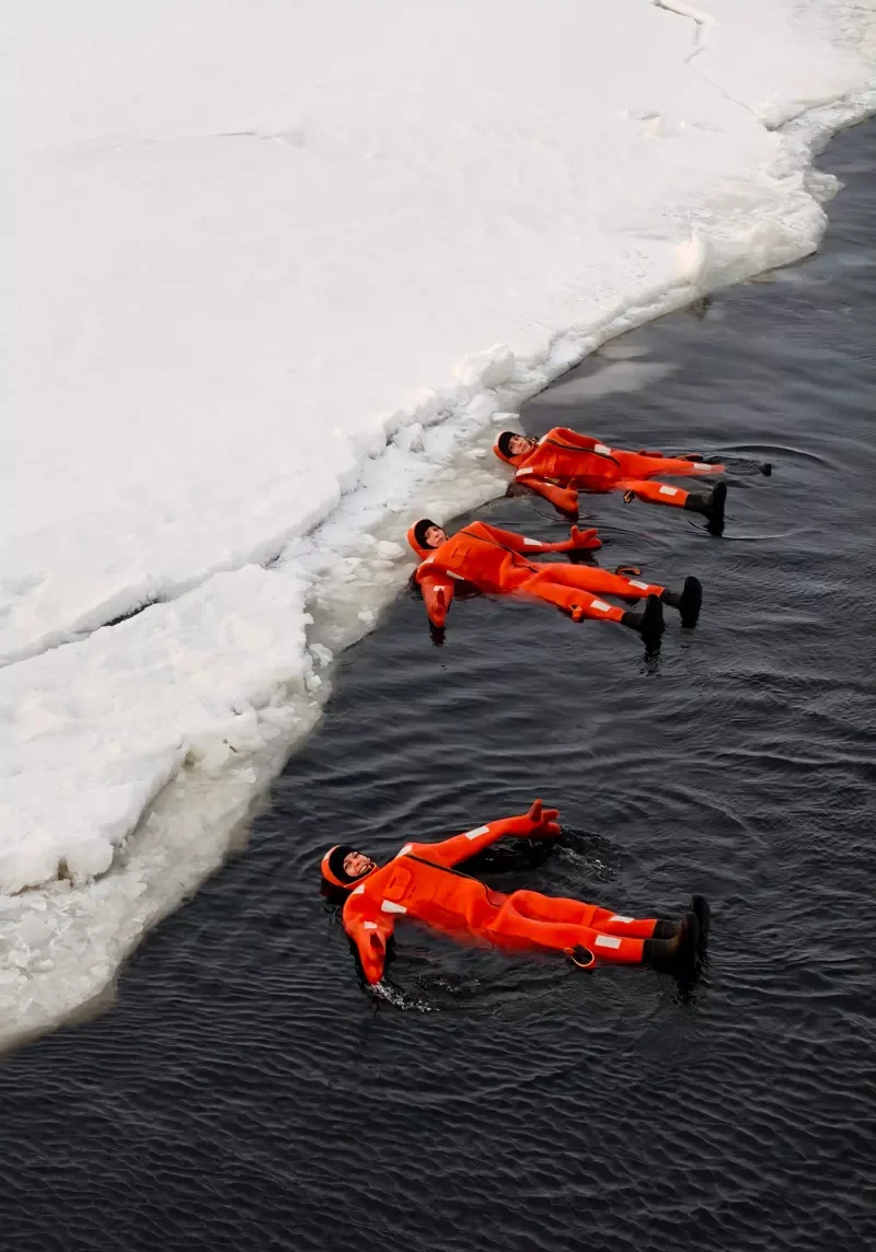 Tourists floating in freezing water near the icy edge of the Bothnian Sea wearing bright orange survival suits during an icebreaker cruise in Finland.