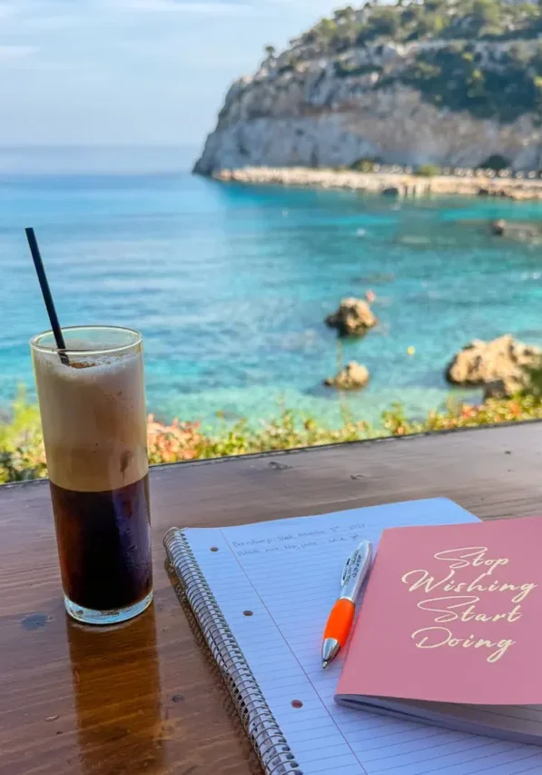 A tall glass of freddo espresso sits beside a pink notebook that says “Stop Wishing Start Doing” and a spiral-bound notepad on a wooden table overlooking the turquoise sea and rocky coastline in Rhodes.