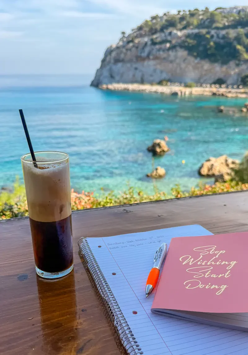 A tall glass of freddo espresso sits beside a pink notebook that says “Stop Wishing Start Doing” and a spiral-bound notepad on a wooden table overlooking the turquoise sea and rocky coastline in Rhodes.