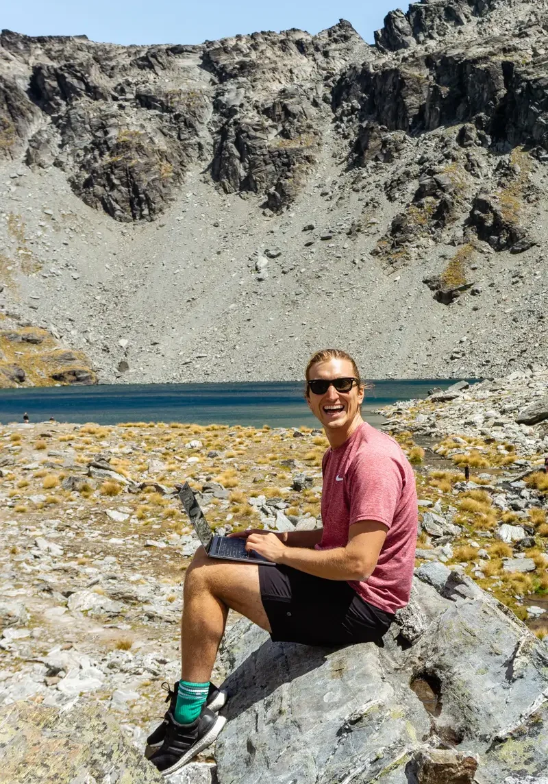 Dom working on his laptop in front of Lake Alta, a beautiful mountain lake near Queenstown in New Zealand.