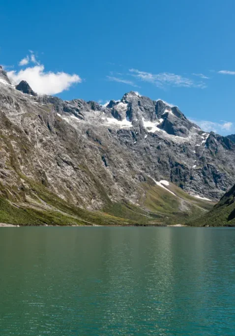Lake Marian, a beautiful alpine lake in Fiordland National Park, is surrounded by steep rocky mountains with patches of snow and lush green vegetation. The calm green lake sits in a remote alpine valley.