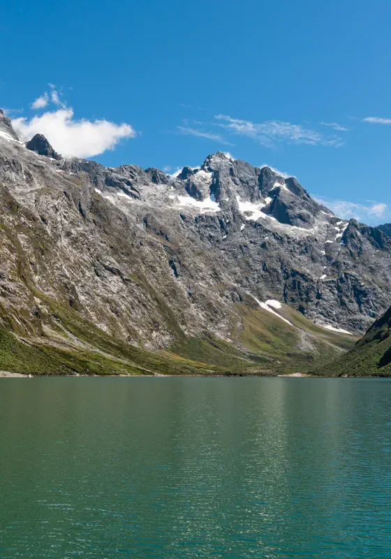 Lake Marian, a beautiful alpine lake in Fiordland National Park, is surrounded by steep rocky mountains with patches of snow and lush green vegetation. The calm green lake sits in a remote alpine valley.