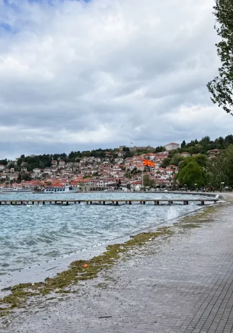 Lakeside promenade in Ohrid with waves crashing along the shore and a hillside town covered in red-roofed buildings under cloudy skies. A large North Macedonian flag waves above the waterfront near the centre of town.