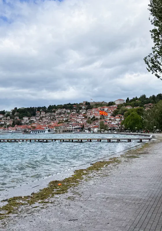 Lakeside promenade in Ohrid with waves crashing along the shore and a hillside town covered in red-roofed buildings under cloudy skies. A large North Macedonian flag waves above the waterfront near the centre of town.