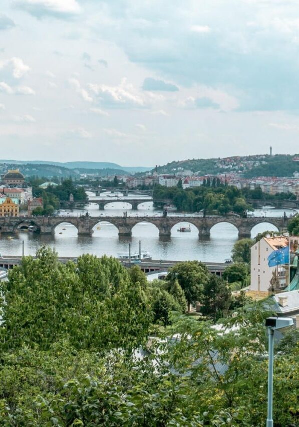 A panoramic view of Prague showcasing the Vltava River lined with historic stone bridges, including the iconic Charles Bridge. The city’s red rooftops, green copper domes, and tree-covered riverbanks frame the water.