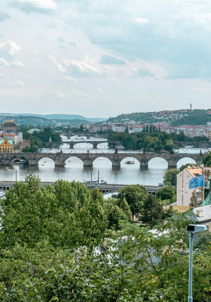 A panoramic view of Prague showcasing the Vltava River lined with historic stone bridges, including the iconic Charles Bridge. The city’s red rooftops, green copper domes, and tree-covered riverbanks frame the water.