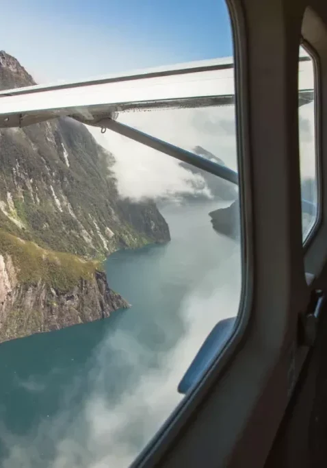 View from a small aircraft window showing a breathtaking aerial perspective of Milford Sound, with steep cliffs rising dramatically from the fiord. A passenger is seen gazing out, capturing the scene on a camera.