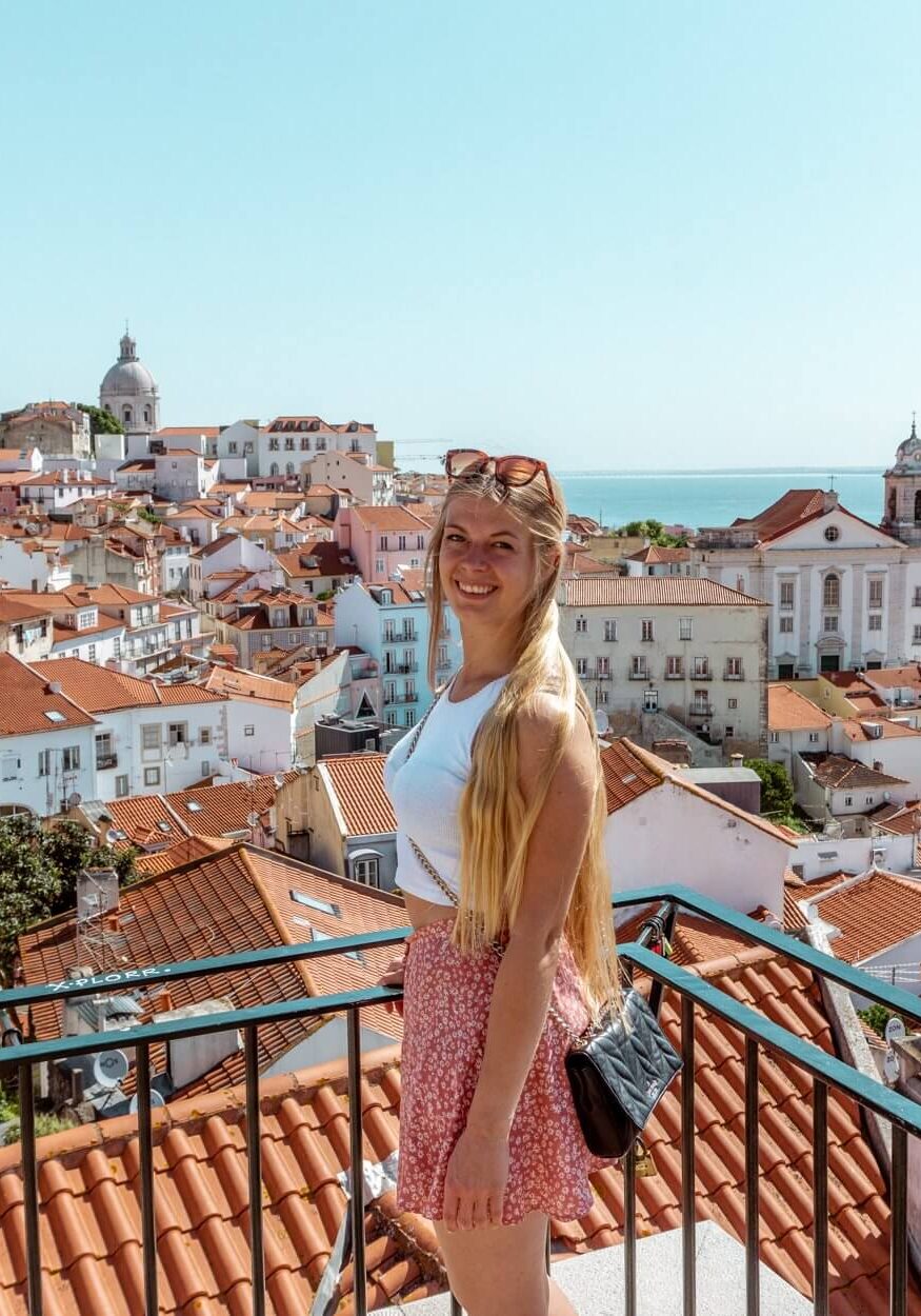 A woman stands at a viewpoint overlooking a historic city neighborhood with red-tiled rooftops and pastel buildings, smiling toward the camera on a sunny day. A large body of water and church domes are visible in the distance beyond the rooftops.
