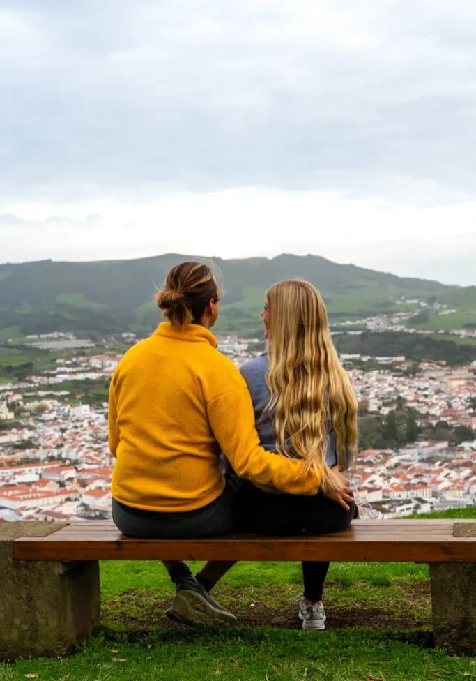 Two people sit side by side on a wooden bench at a hilltop viewpoint on Terceira Island, one with an arm around the other, looking out over the whitewashed rooftops and terracotta tiles of Angra do Heroísmo with green volcanic hills rising behind the city.