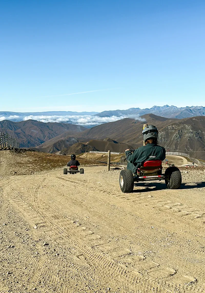 Two riders drive mountain carts downhill on a wide gravel track with sweeping alpine views in the background. The scene highlights the open landscape and sense of adventure at a high-altitude mountain resort in New Zealand.