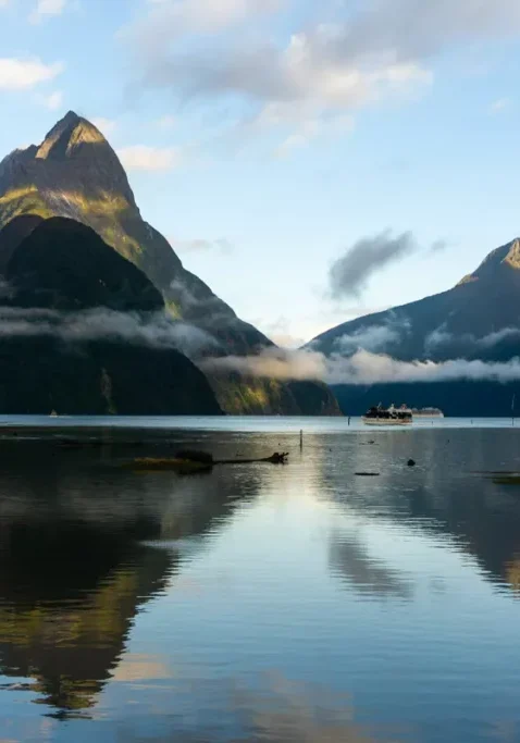 Mitre Peak and the surrounding mountains reflect in the calm water of Milford Sound, New Zealand. Low clouds cling to the slopes, and a small cruise boat crosses the fiord in the distance.