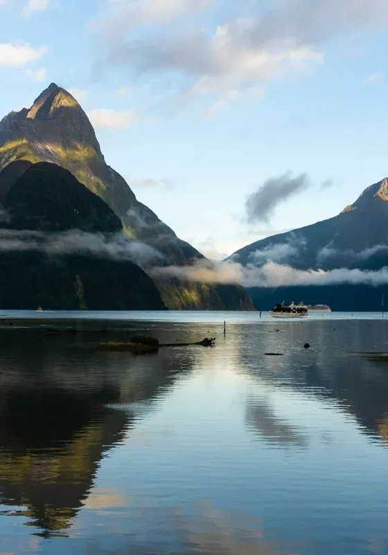Mitre Peak and the surrounding mountains reflect in the calm water of Milford Sound, New Zealand. Low clouds cling to the slopes, and a small cruise boat crosses the fiord in the distance.