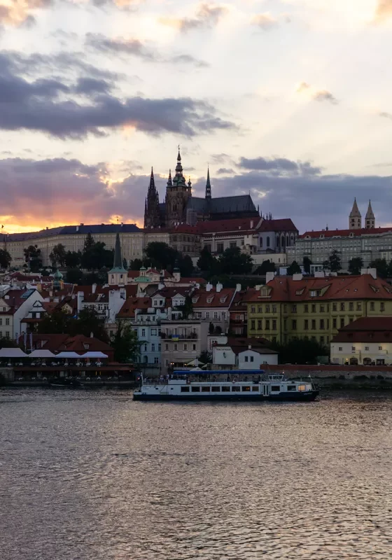 A serene evening view of Prague Castle and St. Vitus Cathedral silhouetted against a dramatic sunset sky, with golden and purple hues peeking through scattered clouds. Below, a boat glides along the Vltava River, passing charming red-roofed buildings lining the riverbank.