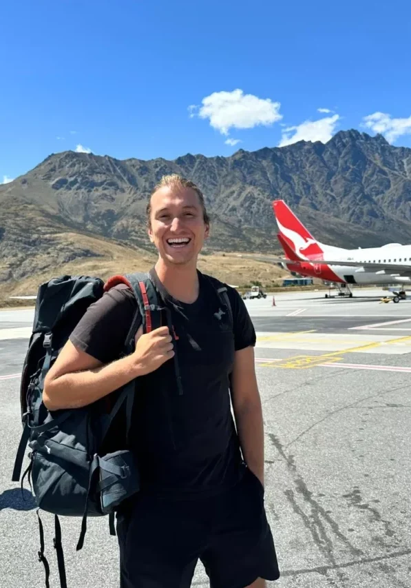 Smiling traveller standing on the tarmac at Queenstown Airport with a large backpack, wearing a black outfit, with a Qantas airplane and the Remarkables mountain range behind him under a sunny sky.