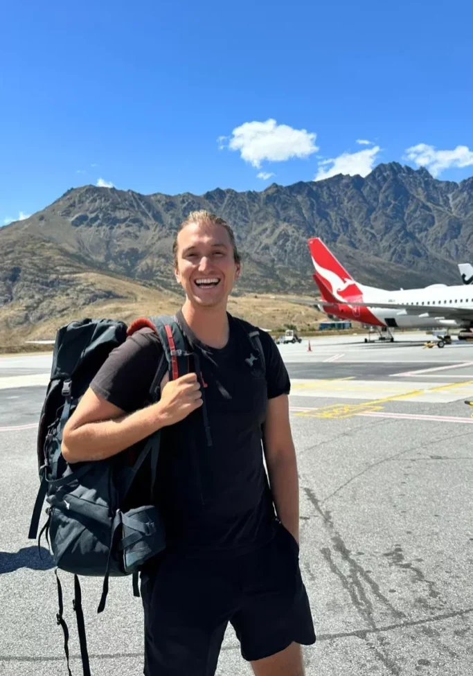 Smiling traveller standing on the tarmac at Queenstown Airport with a large backpack, wearing a black outfit, with a Qantas airplane and the Remarkables mountain range behind him under a sunny sky.