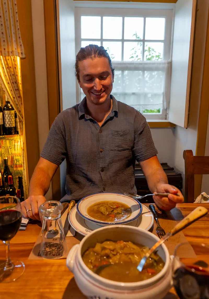 Man smiling while seated at a wooden dining table with a bowl of traditional Azorean soup and a glass of red wine in a cosy restaurant.
