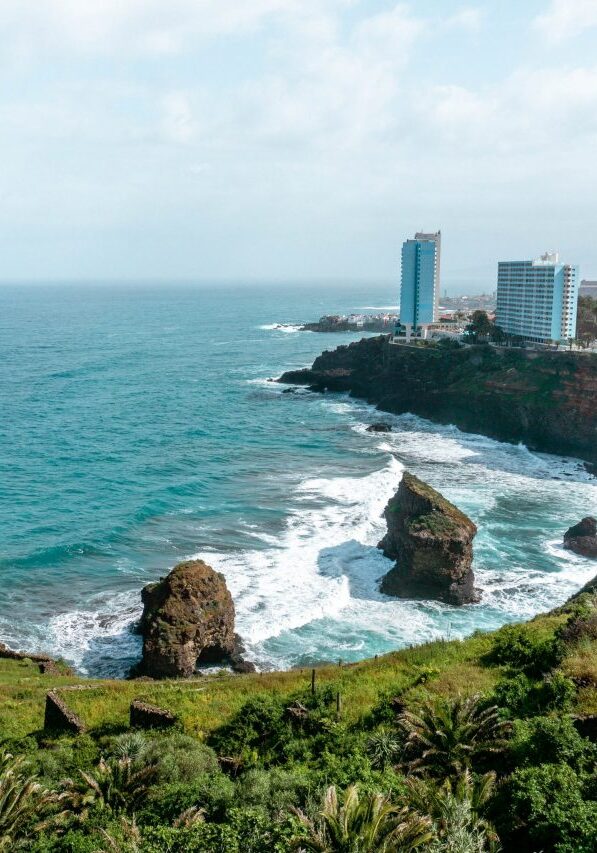 A rugged coastline of Rambla de Castro in Tenerife, Spain, with turquoise waves crashing against rocky sea stacks and cliffs. High-rise buildings overlook the Atlantic Ocean from the lush, green hillside, blending natural beauty with urban development.
