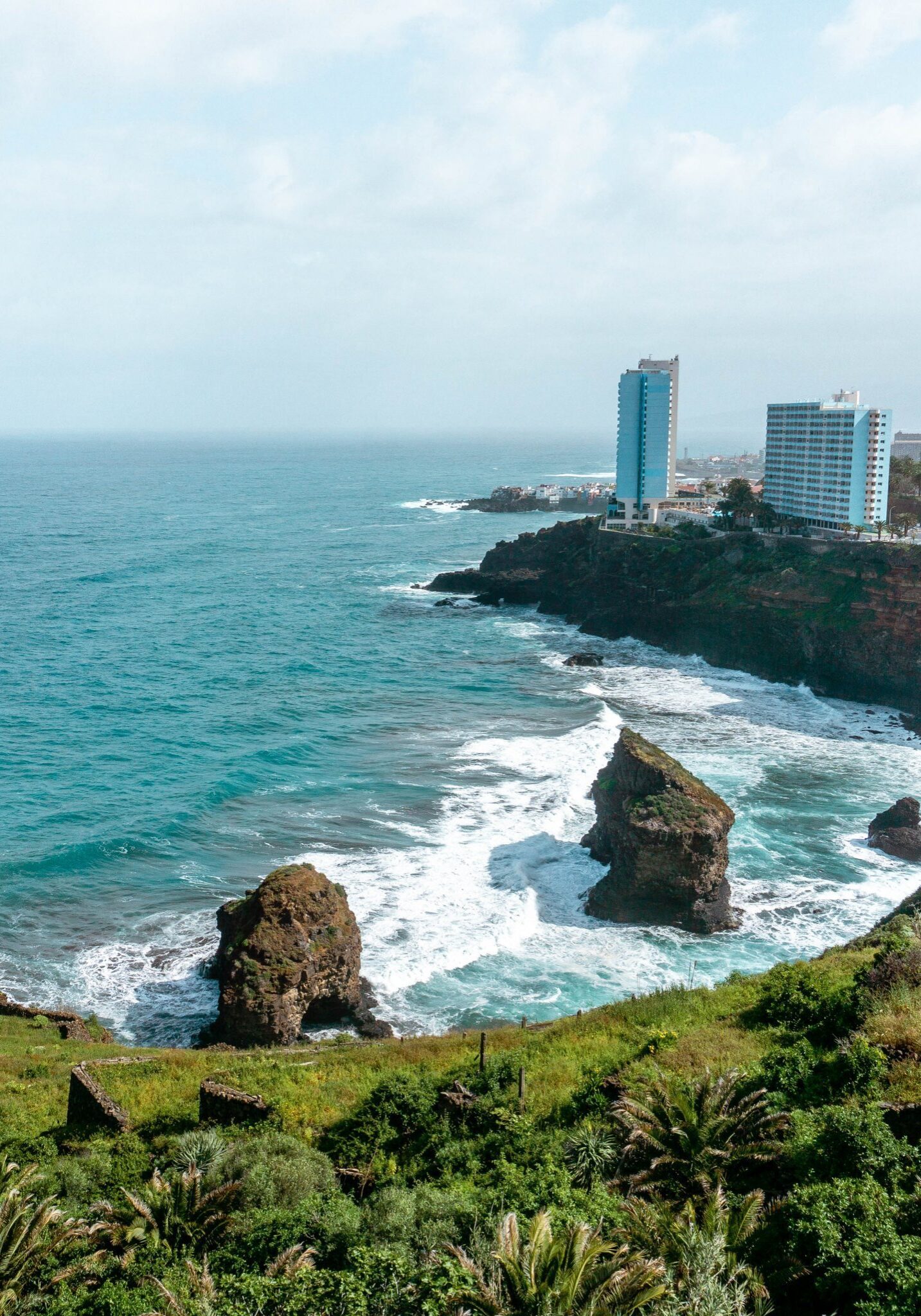 A rugged coastline of Rambla de Castro in Tenerife, Spain, with turquoise waves crashing against rocky sea stacks and cliffs. High-rise buildings overlook the Atlantic Ocean from the lush, green hillside, blending natural beauty with urban development.