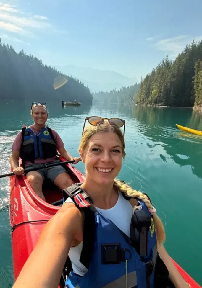 Two people kayaking on a turquoise river surrounded by forested mountains, with the woman taking a selfie from the front of the red kayak while both wear life jackets.