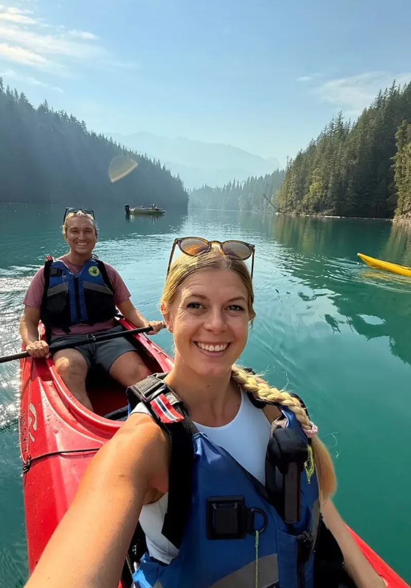 Two people kayaking on a turquoise river surrounded by forested mountains, with the woman taking a selfie from the front of the red kayak while both wear life jackets.